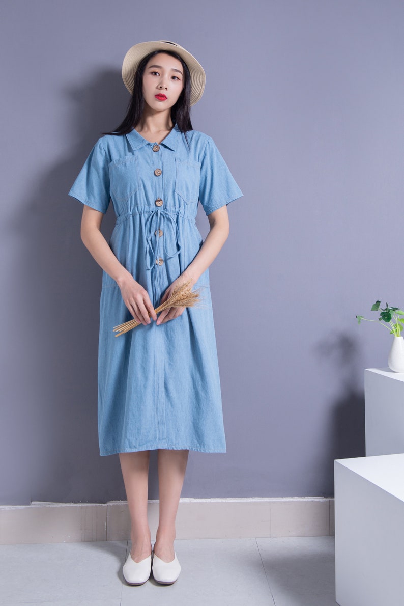 May include: A woman wearing a light blue denim dress with a straw hat and white shoes. She is holding a bunch of dried wheat stalks.