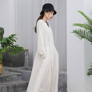Puede incluir: Un vestido largo color crema con mangas largas y botones en la parte delantera. El vestido tiene un cintur&oacute;n anudado en la cintura y bolsillos. Un sombrero de pescador negro y zapatos blancos completan el conjunto. El vestido es de tela ligera.