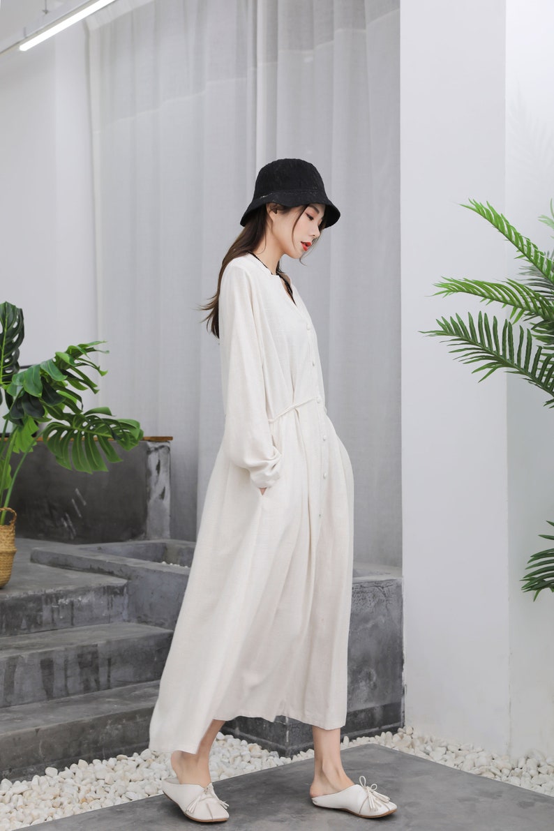 Puede incluir: Un vestido largo color crema con mangas largas y botones en la parte delantera. El vestido tiene un cintur&oacute;n anudado en la cintura y bolsillos. Un sombrero de pescador negro y zapatos blancos completan el conjunto. El vestido es de tela ligera.