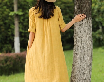 May include: A woman wearing a yellow linen dress and a straw hat with a black and white checkered band. The dress has short sleeves and pockets. The person is standing near a tree in a grassy area.