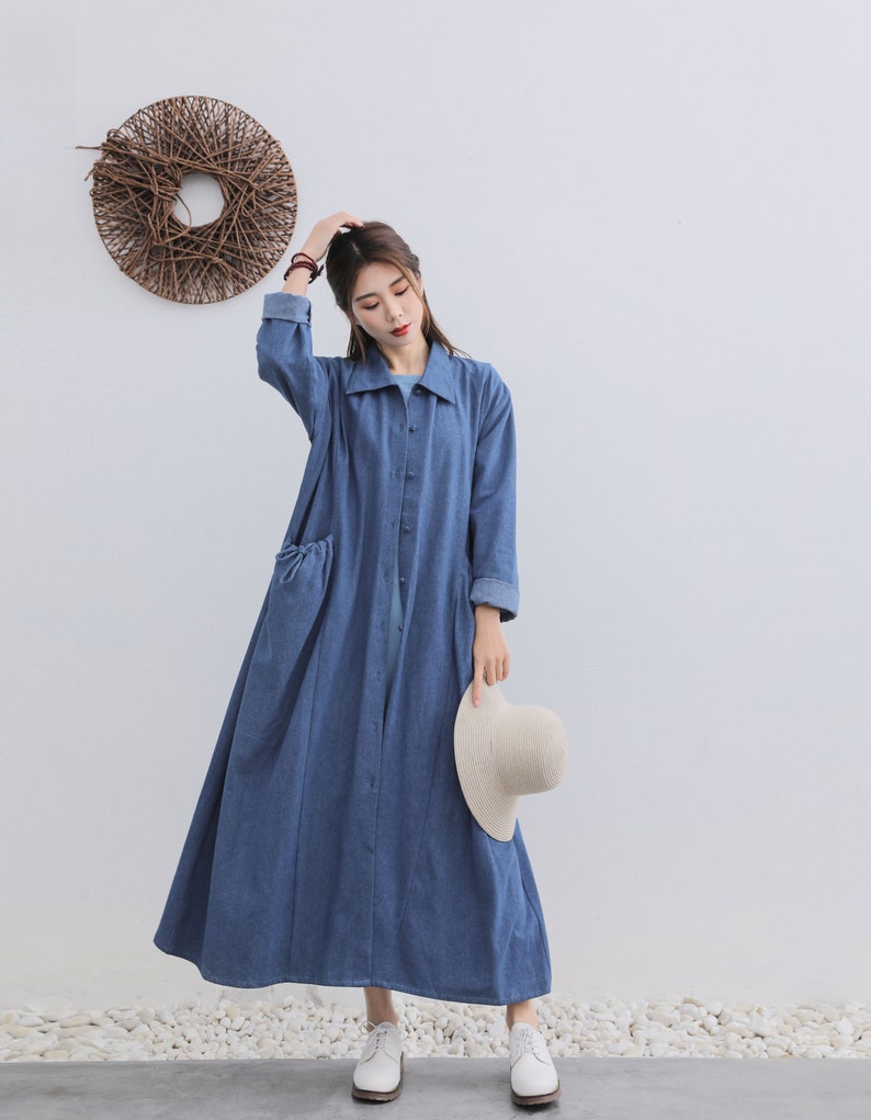 May include: A long, blue denim dress with a collared neckline and button fastening. The dress has a relaxed fit and features two pockets on the front. The model is wearing white shoes and holding a straw hat.