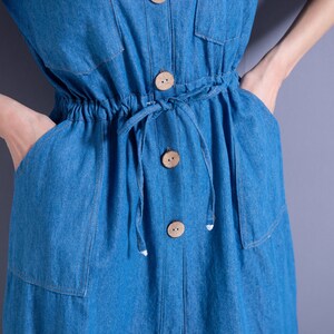 May include: A blue denim dress with a drawstring waist and wooden buttons. The dress has two pockets on the front.