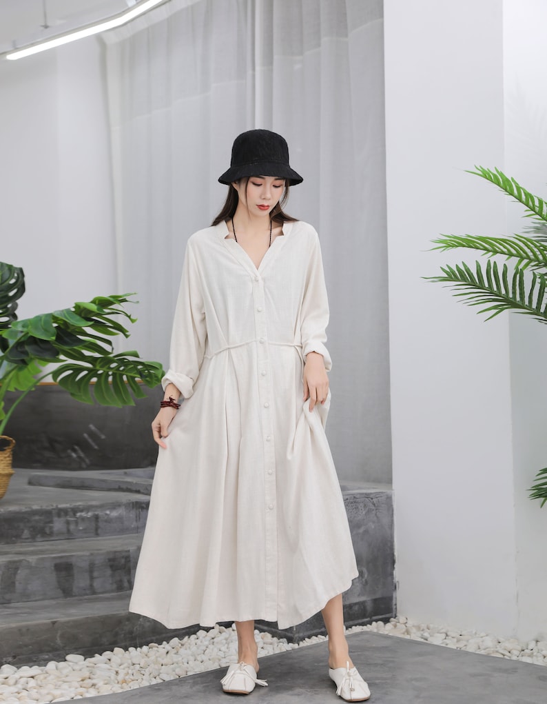 Puede incluir: Vestido largo beige claro de manga larga, abotonado y con cuello en V. El vestido tiene cintura ce&ntilde;ida y falda fluida. Un sombrero de pescador negro y zapatos blancos completan el conjunto. El vestido es de tejido ligero.