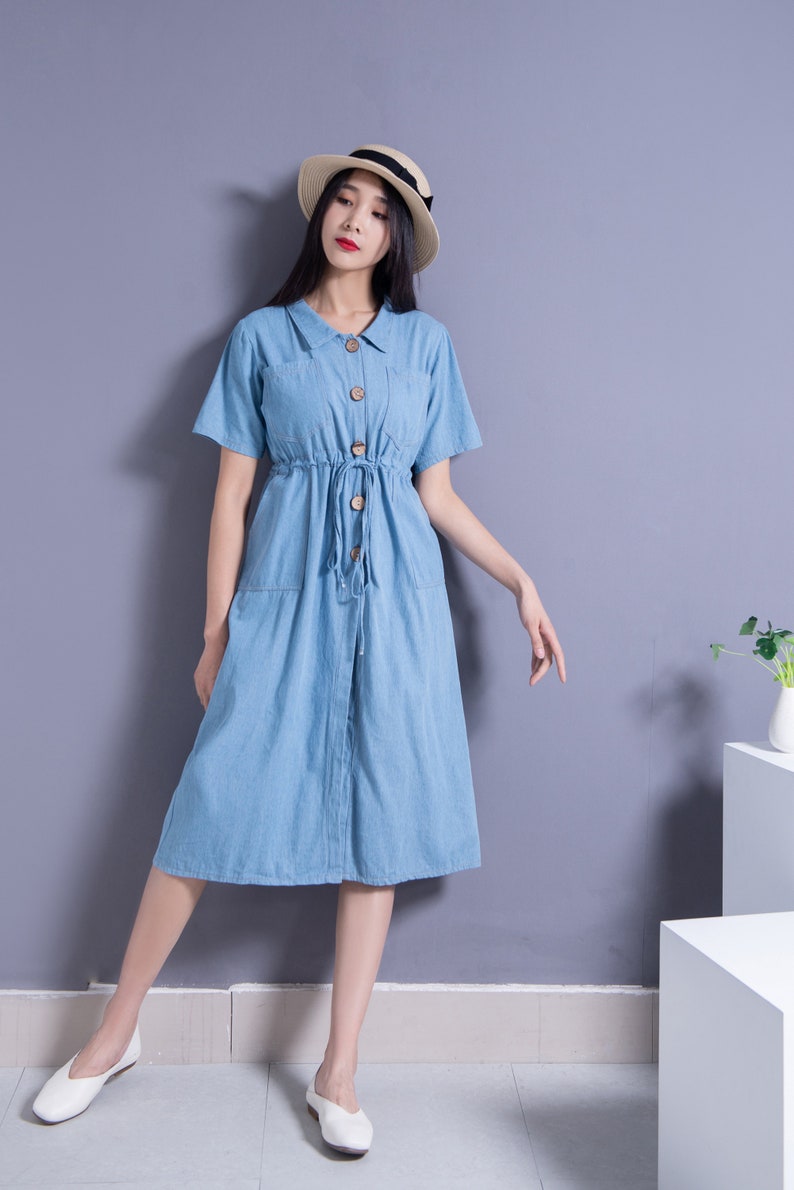 May include: A woman wearing a light blue denim dress with a button-up front and a drawstring waist. She is wearing a straw hat with a black ribbon and white flats.