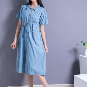 May include: A woman wearing a light blue denim dress with a drawstring waist and short sleeves. She is wearing a straw hat with a black ribbon and white flats. The dress has a button-up front and two pockets.