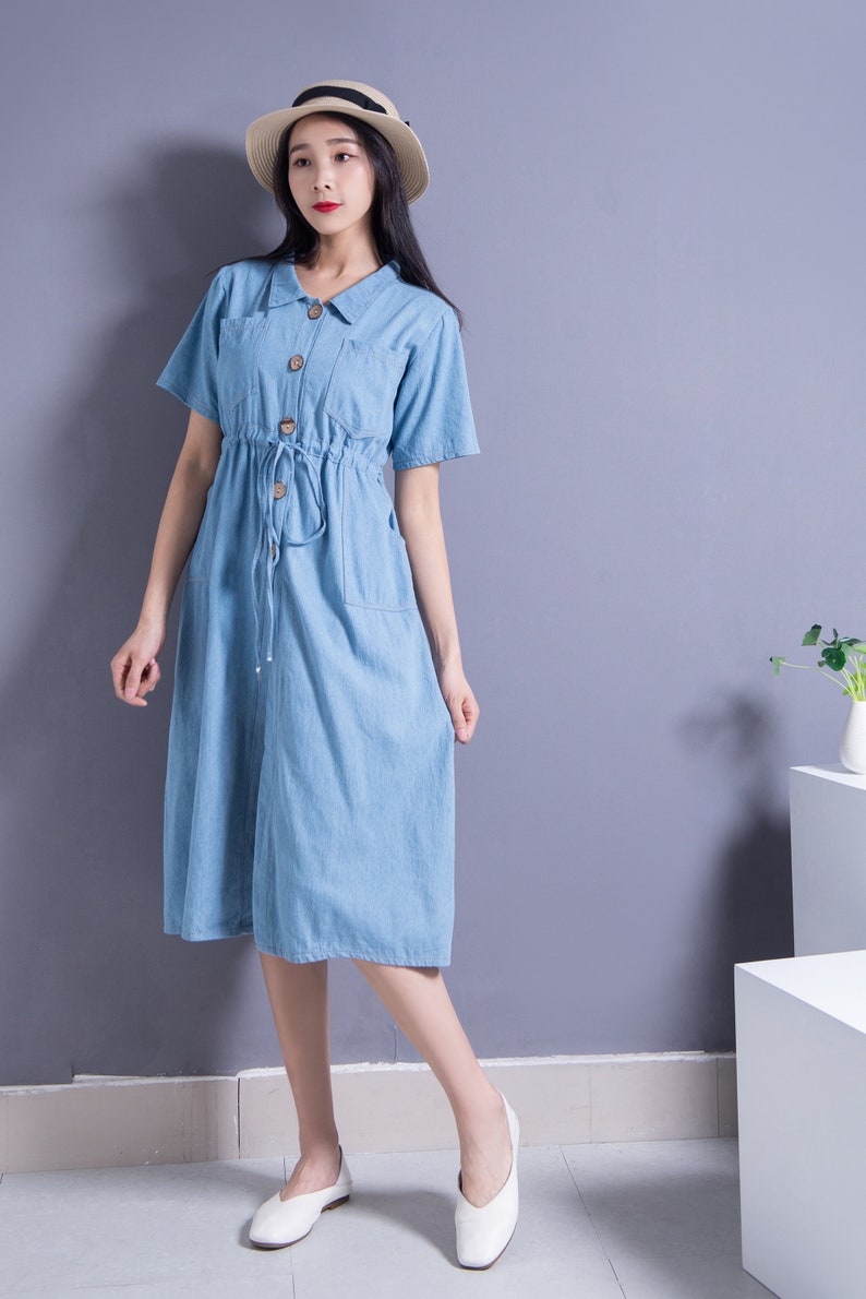 May include: A woman wearing a light blue denim dress with a drawstring waist and short sleeves. She is wearing a straw hat with a black ribbon and white flats. The dress has a button-up front and two pockets.