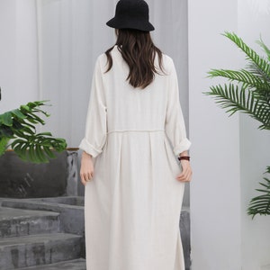Puede incluir: Un vestido largo color crema con mangas largas y falda plisada. El vestido se lleva con un sombrero de pescador negro. La modelo est&aacute; de pie frente a una pared blanca con una escalera y vegetaci&oacute;n.