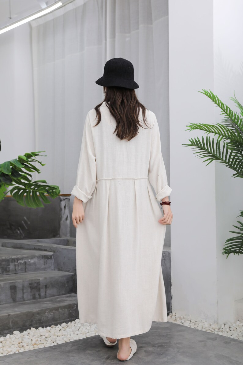 Puede incluir: Un vestido largo color crema con mangas largas y falda plisada. El vestido se lleva con un sombrero de pescador negro. La modelo est&aacute; de pie frente a una pared blanca con una escalera y vegetaci&oacute;n.