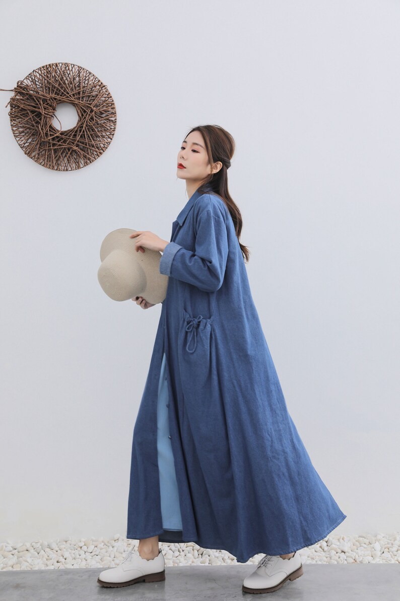 May include: A long, blue denim shirt dress with a button-up front and a tie at the waist. The dress has a large pocket on the left side and is worn with white shoes.