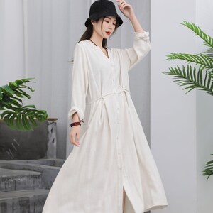 Puede incluir: Vestido largo color crema con botones, cuello en V y mangas largas. Tiene un lazo en la cintura y se lleva con un sombrero negro y zapatos blancos. El vestido est&aacute; hecho de una tela ligera, ideal para el verano.