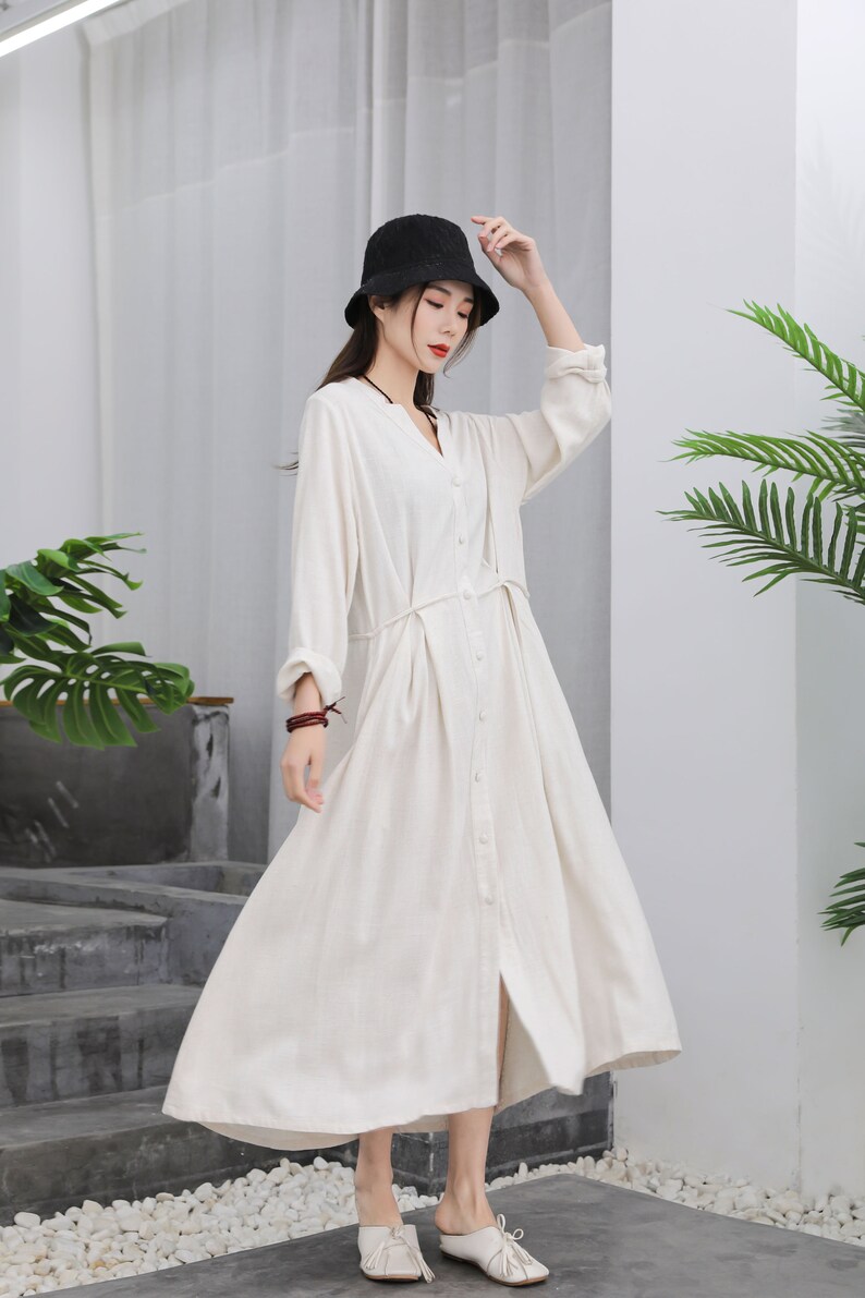 Puede incluir: Vestido largo color crema con botones, cuello en V y mangas largas. Tiene un lazo en la cintura y se lleva con un sombrero negro y zapatos blancos. El vestido est&aacute; hecho de una tela ligera, ideal para el verano.