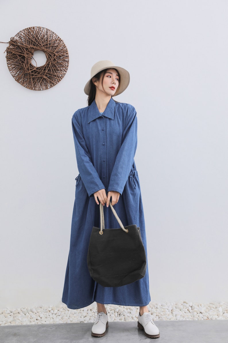May include: A woman wearing a blue denim shirt dress with a white hat and a black tote bag with rope handles. She is standing on a white stone surface.