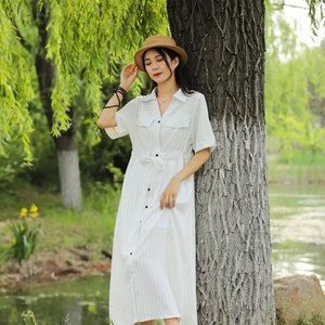 May include: A woman wearing a white striped dress with a brown hat stands next to a tree by a pond.