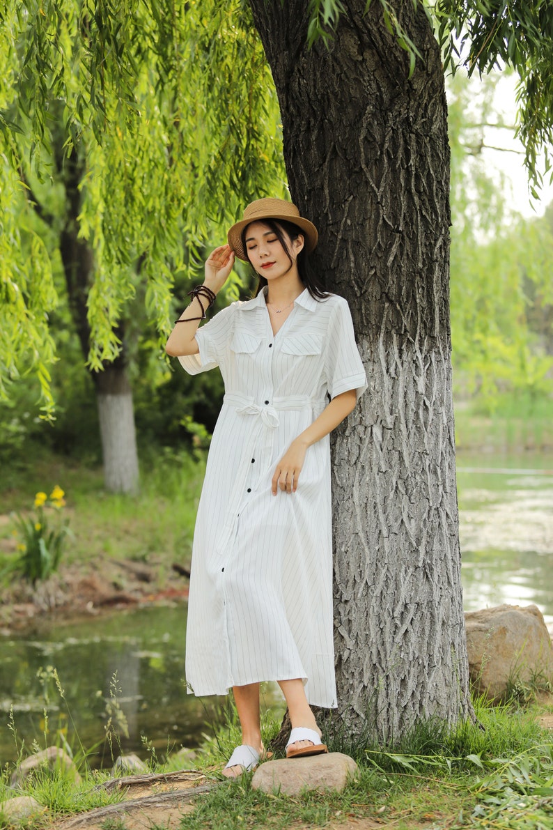 May include: A woman wearing a white striped dress with a brown hat stands next to a tree by a pond.