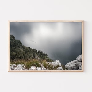 Op de afbeelding: Een panoramisch uitzicht vanaf een bergtop met uitzicht op een uitgestrekt wateroppervlak. Het water is donkergrijs en de lucht is lichtgrijs met een paar wolken. De berghelling is bedekt met groene bomen en rotsen.