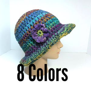 May include: A multicolored crochet bucket hat featuring a textured yarn pattern in shades of blue, green, purple, and yellow. The hat includes a prominent purple crocheted flower accent on the side and a short brim. The text 8 Colors is visible.