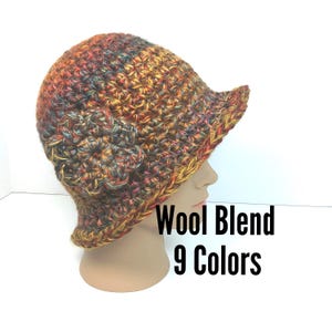 May include: A crocheted bucket hat made from a variegated wool blend yarn in shades of brown, orange, yellow, and dark red. The hat features a large, dimensional crocheted flower on the side and includes the text Wool Blend 9 Colors.