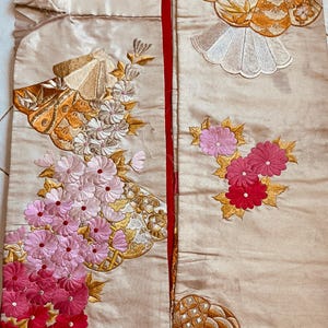 Op de afbeelding: Cr&egrave;mekleurige stof met ingewikkelde borduursels met bloemen- en waaiermotieven. Het borduurwerk bevat tinten roze, rood, goud en wit. De stof is verdeeld door een rode naad, wat wijst op een kledingstuk of textielstuk.