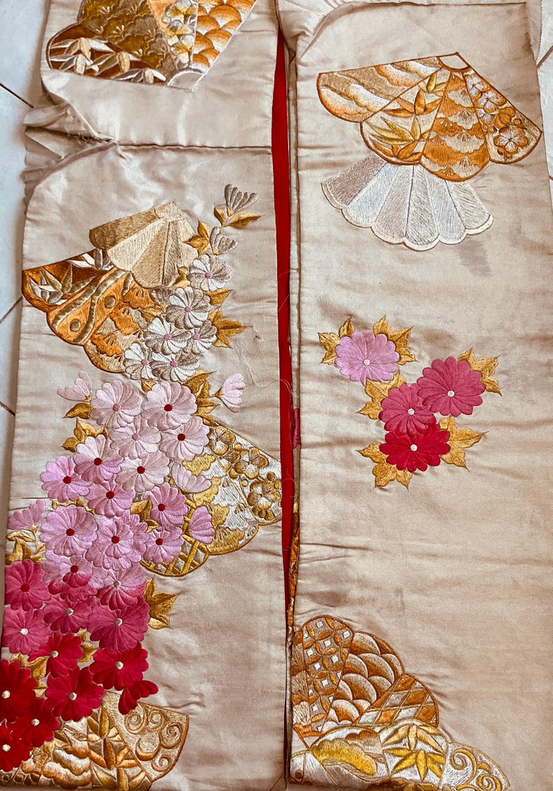 Op de afbeelding: Cr&egrave;mekleurige stof met ingewikkelde borduursels met bloemen- en waaiermotieven. Het borduurwerk bevat tinten roze, rood, goud en wit. De stof is verdeeld door een rode naad, wat wijst op een kledingstuk of textielstuk.