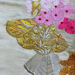 Op de afbeelding: Close-up van een geborduurd textiel met bloemen- en geometrische patronen. De borduursels tonen een vlindervorm met gouden, gele en zilveren details, naast roze en oranje bloemen. De achtergrond is lichtbeige.