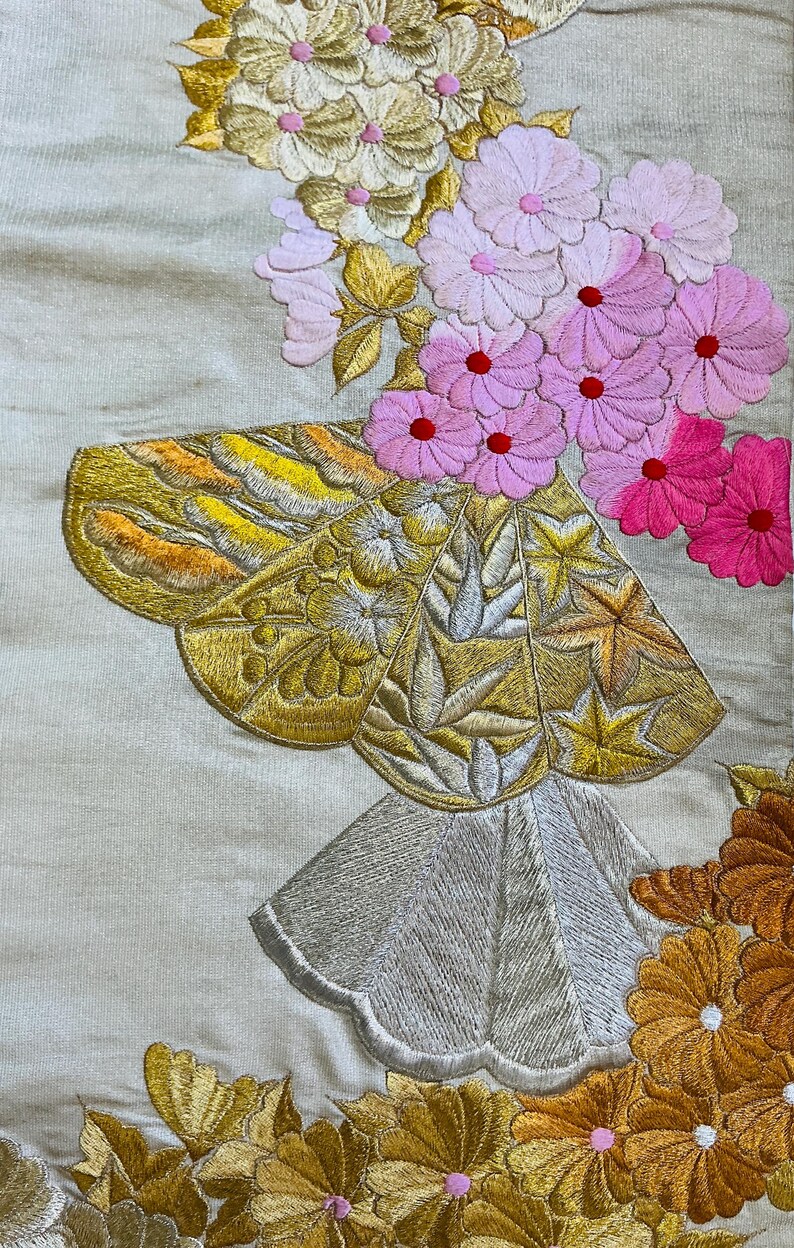 Op de afbeelding: Close-up van een geborduurd textiel met bloemen- en geometrische patronen. De borduursels tonen een vlindervorm met gouden, gele en zilveren details, naast roze en oranje bloemen. De achtergrond is lichtbeige.