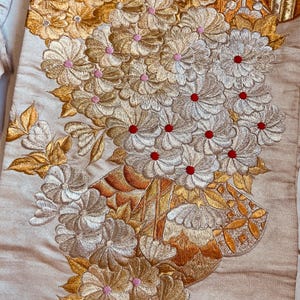 Op de afbeelding: Close-up van een beige textiel met ingewikkelde bloemenborduurwerk. Het ontwerp toont clusters van bloemen in goud, zilver en roze, met rode centra. Het borduurwerk is gedetailleerd en sierlijk, wat een textuur en elegante uitstraling cre&euml;ert.