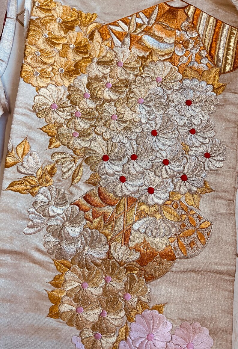 Op de afbeelding: Close-up van een beige textiel met ingewikkelde bloemenborduurwerk. Het ontwerp toont clusters van bloemen in goud, zilver en roze, met rode centra. Het borduurwerk is gedetailleerd en sierlijk, wat een textuur en elegante uitstraling cre&euml;ert.