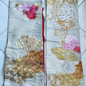 Op de afbeelding: Twee cr&egrave;mekleurige, geborduurde stoffen panelen. E&eacute;n paneel heeft een cluster van roze en rode bloemen, de andere heeft bloemen in wit, goud en roze. Beide panelen hebben waaiervormige ontwerpen. Waarschijnlijk onderdeel van een kledingstuk.