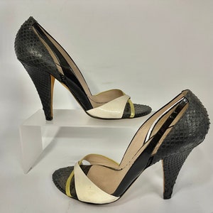 May include: A pair of high-heeled shoes with a snakeskin pattern on the heel and toe. The shoes have a black and white color scheme with a gold accent on the toe strap. The heels are approximately 4 inches tall.