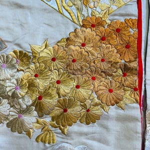 Op de afbeelding: Detail van een textiel met ingewikkelde borduursels. Het ontwerp toont clusters van goudgele bloemen met rode centra, naast een waaier motief. De achtergrond is lichtbeige, met een rode rand aan de rechterkant.