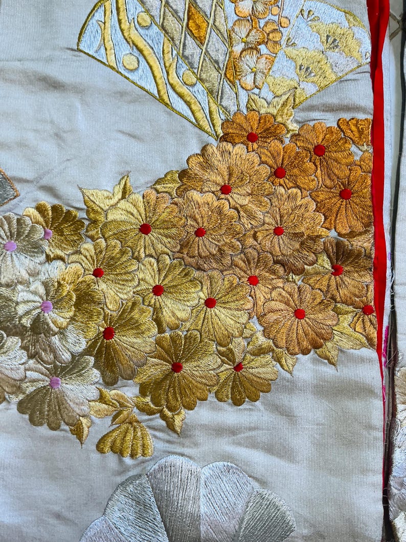Op de afbeelding: Detail van een textiel met ingewikkelde borduursels. Het ontwerp toont clusters van goudgele bloemen met rode centra, naast een waaier motief. De achtergrond is lichtbeige, met een rode rand aan de rechterkant.