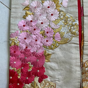 Op de afbeelding: Close-up van geborduurde stof met bloemmotieven. De stof is lichtbeige, met roze en rode bloemenborduurwerk. Gouden en witte borduursels accentueren het ontwerp. Een rode rand is zichtbaar.