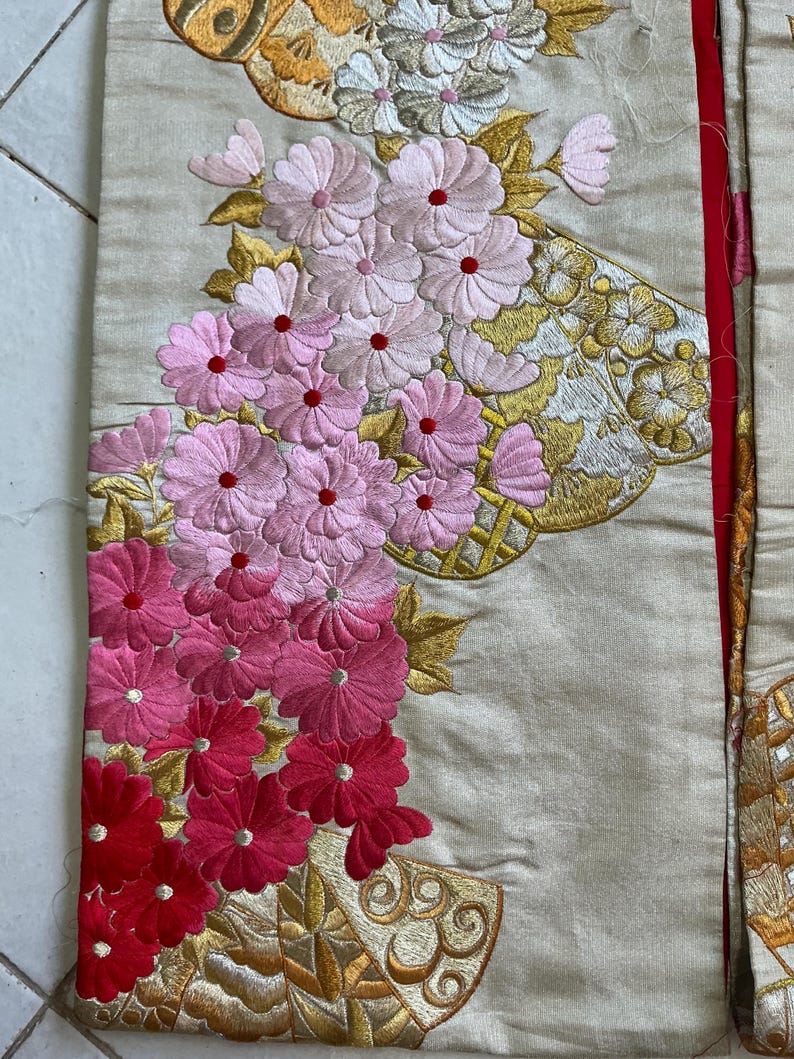 Op de afbeelding: Close-up van geborduurde stof met bloemmotieven. De stof is lichtbeige, met roze en rode bloemenborduurwerk. Gouden en witte borduursels accentueren het ontwerp. Een rode rand is zichtbaar.