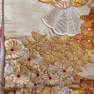 Op de afbeelding: Close-up van een beige textiel met ingewikkelde gouden en zilveren borduursels. Het ontwerp toont gedetailleerde bloempatronen met rode en roze accenten en een gestileerde figuur met een gouden halo.