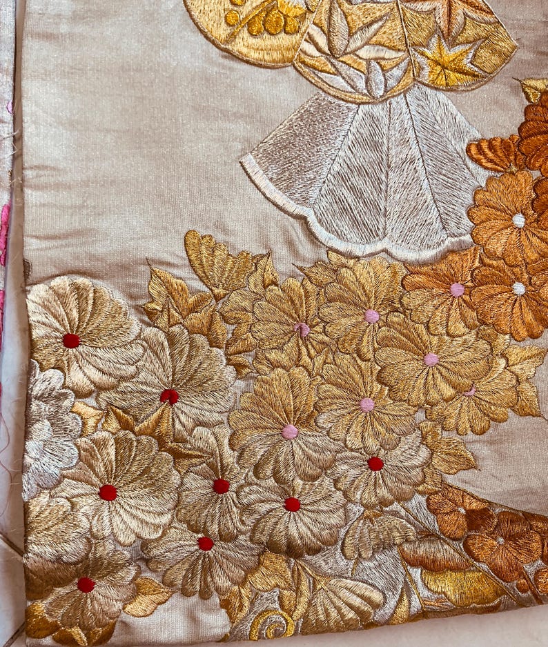 Op de afbeelding: Close-up van een beige textiel met ingewikkelde gouden en zilveren borduursels. Het ontwerp toont gedetailleerde bloempatronen met rode en roze accenten en een gestileerde figuur met een gouden halo.