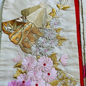 Op de afbeelding: Close-up van een cr&egrave;mekleurig textiel met ingewikkelde borduursels. Het ontwerp bevat gouden en bruine vlinder- en bloemmotieven, waaronder roze en witte bloemen met rode centra. Een rode streep loopt langs de rechterrand.