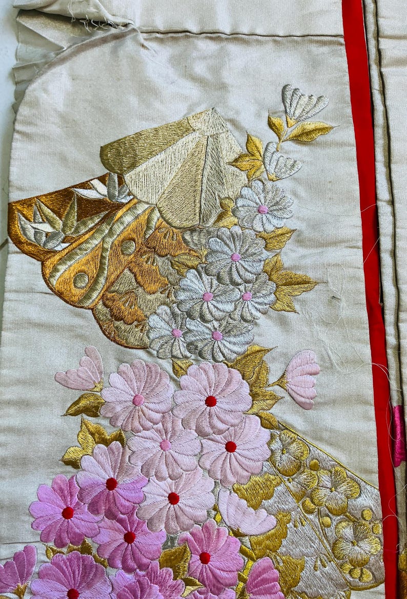 Op de afbeelding: Close-up van een cr&egrave;mekleurig textiel met ingewikkelde borduursels. Het ontwerp bevat gouden en bruine vlinder- en bloemmotieven, waaronder roze en witte bloemen met rode centra. Een rode streep loopt langs de rechterrand.