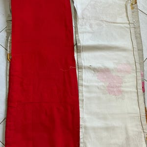 Op de afbeelding: Een felrood stoffen paneel naast een wit stoffen paneel met roze bloempatronen. Het witte paneel heeft een decoratieve rand met gouden accenten. De stoffen liggen op een lichtgekleurd oppervlak.