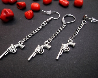 May include: A pair of silver gun charm earrings with a chain and a hook closure. The earrings are hanging from a black surface.