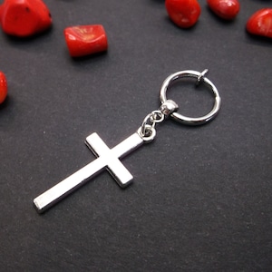 May include: Silver cross charm hanging from a silver hoop earring. The earring is on a black surface with red coral pieces scattered around it.