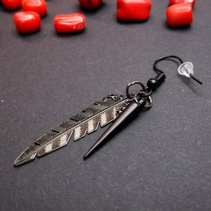 May include: A silver feather earring with a black spike hanging from a black hook. The earring is on a black surface.