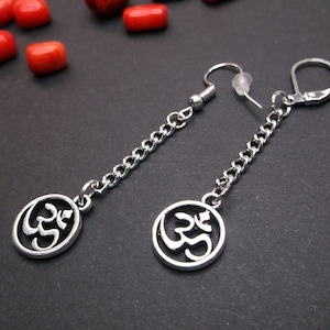 May include: Silver earrings with a circular pendant featuring an Om symbol. The earrings are attached to a silver chain.