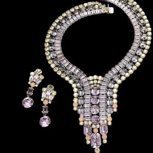 May include: A vintage rhinestone necklace and earrings set. The necklace features a geometric design with a cascading drop. The set is made of clear and light purple rhinestones.