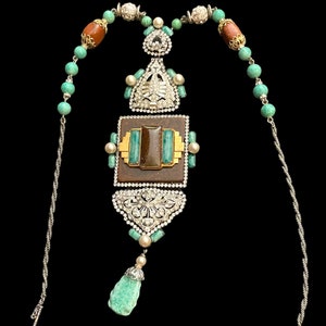 May include: A silver chain necklace with a large pendant featuring a brown and green stone surrounded by smaller green stones and silver accents. The pendant is attached to a silver chain with a turquoise and brown bead strand.