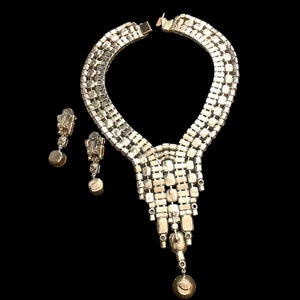 May include: A silver tone rhinestone necklace and earrings set. The necklace features a cascading design with a teardrop pendant. The earrings are clip-on style.