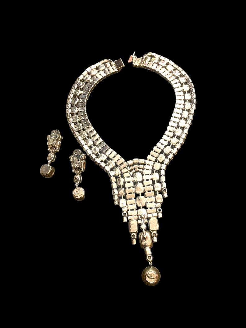 May include: A silver tone rhinestone necklace and earrings set. The necklace features a cascading design with a teardrop pendant. The earrings are clip-on style.
