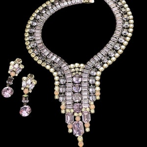 May include: A vintage rhinestone necklace and earrings set with a cascading design. The necklace features a wide band of clear and lavender rhinestones, with a dangling section of lavender and clear rhinestones. The earrings are dangle earrings with a similar design.