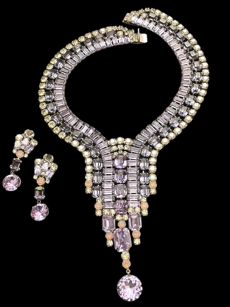 May include: A vintage rhinestone necklace and earrings set with a cascading design. The necklace features a wide band of clear and lavender rhinestones, with a dangling section of lavender and clear rhinestones. The earrings are dangle earrings with a similar design.