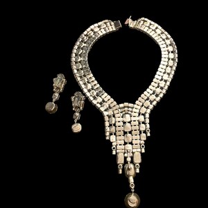 May include: A silver tone necklace with a cascading design of clear rhinestones. The necklace is paired with matching earrings with a single clear rhinestone drop.