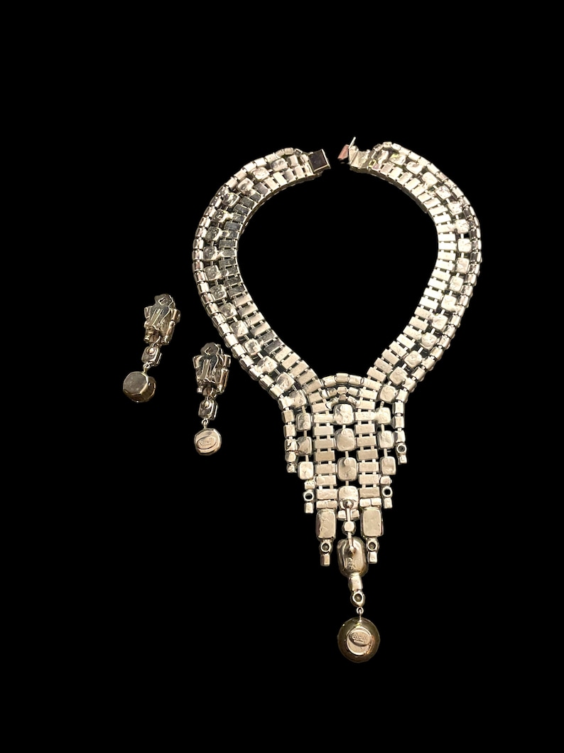 May include: A silver tone necklace with a cascading design of clear rhinestones. The necklace is paired with matching earrings with a single clear rhinestone drop.